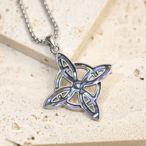 May include: A silver-toned pendant necklace featuring a four-pointed Celtic knot design. The metal frame is inlaid with iridescent abalone shell, displaying shifting hues of blue, green, and purple. The pendant hangs from a matching silver-toned chain.