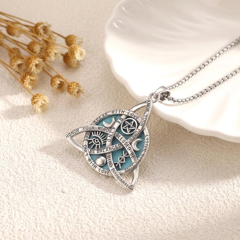 May include: A silver-toned pendant necklace featuring a triquetra shape surrounding a circular light blue inlay. The metal surface is engraved with symbols including a pentagram, crescent moon, eye, and runes, along with the text "SPEAK WITH LOVE".