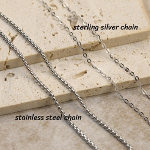 May include: Two silver-coloured metal chains are displayed. On the left, a thicker, rounded stainless steel chain. On the right, a thinner, flat-linked sterling silver chain. Text labels identify each chain as stainless steel chain and sterling silver chain.