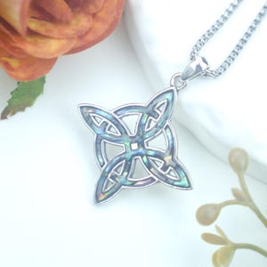 May include: A silver-toned pendant necklace featuring a Celtic knot design with iridescent abalone shell inlay. The pendant is suspended from a delicate chain. The background includes a white surface and a blurred orange flower.