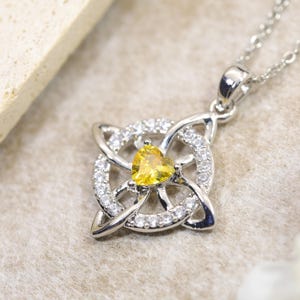 May include: A silver-toned pendant necklace featuring a central heart-shaped yellow gemstone set within a circular frame of clear white stones. The metal frame displays an intricate, interlaced knotwork design, suspended from a delicate silver chain.