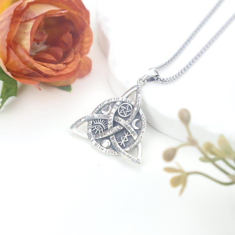 May include: A silver-colored pendant necklace featuring a Celtic knot design. The pendant has an intricate pattern with symbols and text, including phrases like "Speak with Love" and "Honor Your Word". The necklace is on a silver chain.