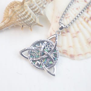 May include: A silver-toned pendant necklace featuring a triquetra frame surrounding a circular iridescent abalone shell inlay. The metal surface is engraved with symbols including a pentagram, crescent moon, and eye, along with the text: SPEAK WITH LOVE, BE KIND, BLESSED BE, BE BALANCED, FIND BOUNDARIES, LISTEN WITH LOVE, KNOW YOUR POWER, HONOR YOUR WORD, SEE ONLY WITH LOVE.
