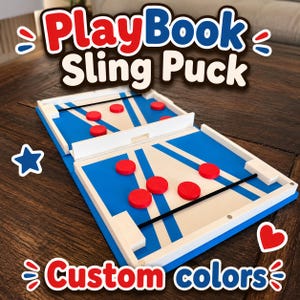 Pode incluir: Um jogo de mesa de disco com elástico, apresentando um tabuleiro azul e branco com um portão central e elásticos pretos. O jogo inclui vários discos circulares vermelhos posicionados em ambos os lados. O texto PlayBook Sling Puck e custom colors é visível.