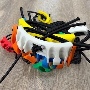 Op de afbeelding: Een verzameling veelkleurige kunststof onderdelen met gebogen, gegroefde randen in wit, geel, oranje, rood, blauw en groen. Elk onderdeel is met elkaar verbonden door dunne zwarte elastische koorden, wat een verwarde, gelaagde structuur vormt.