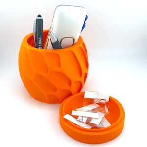 Peut inclure: Un organiseur de bureau orange imprimé en 3D avec un design texturé et organique. Il contient des stylos, un téléphone et d'autres petits objets. À côté, un petit récipient rond orange avec un couvercle, rempli de petites étiquettes blanches.