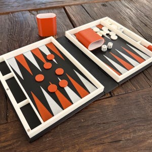Könnte beinhalten: Ein kompaktes Backgammon-Set mit schwarzer Spielfläche, orangefarbenen und weißen Dreiecksmarkierungen und orangefarbenen Spielsteinen. Enthält zwei orangefarbene Würfelbecher, Würfel und weiße Spielsteine. Das Set ist auf einer Holzoberfläche geöffnet.