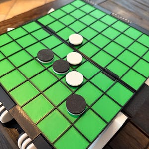 Può includere: Un gioco da tavolo pieghevole con un tabellone verde a griglia e bordi neri. Il gioco include pedine circolari nere e bianche. Il tabellone è appoggiato su una superficie di legno scuro.
