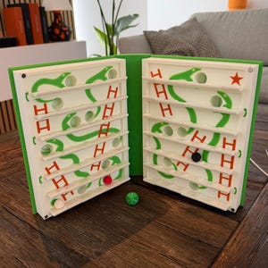 Könnte beinhalten: Ein Schlangen- und Leitern-Brettspiel mit grünem Rand. Es zeigt weiße Spielbretter mit grünen Schlangenillustrationen und roten Leiterillustrationen. Enthält einen grünen Würfel und farbige Spielfiguren.