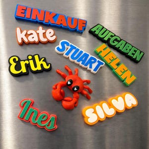 Puede incluir: Una colección de imanes de plástico impresos en 3D de colores vivos con las palabras EINKAUF, AUFGABEN, KATE, STUART, HELEN, ERIK, INES y SILVA, junto a una pequeña figura de cangrejo rojo con ojos en forma de corazón sobre una superficie metálica.