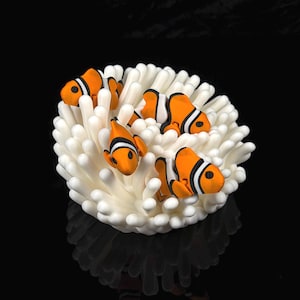 Könnte beinhalten: Eine dekorative Skulptur mit drei orange-weißen Clownfischen, die in einem Bündel aus weißen, abgerundeten, fingerartigen Korallenstrukturen sitzen. Die Fische haben markante schwarze Streifen und sind auf verschiedenen Ebenen angeordnet.