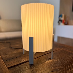 Può includere: Una lampada da tavolo cilindrica con paralume a coste bianco sporco. La lampada è sostenuta da tre gambe rettangolari grigie. La lampada è su un tavolo di legno marrone scuro.