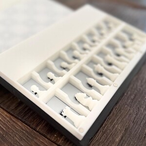 Puede incluir: Un juego de ajedrez blanco con borde negro, cuidadosamente dispuesto en una caja dividida. Las piezas est&aacute;n en sus posiciones iniciales, listas para jugar. El juego est&aacute; sobre una superficie de madera.