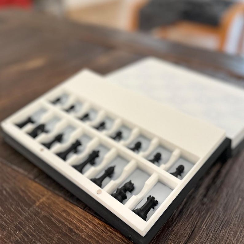 Puede incluir: Un juego de ajedrez en blanco y negro con todas las piezas cuidadosamente dispuestas en una caja dividida. La caja est&aacute; abierta, revelando las piezas de ajedrez en sus compartimentos individuales. Las piezas son negras y la caja es blanca y negra.