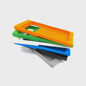 Op de afbeelding: Een stapel kleurrijke rechthoekige telefoonhoesjes in oranje, groen, wit, blauw, zwart en grijs. Op de bovenste oranje hoes staat de tekst "www.1fer.de". De hoesjes zijn onder een lichte hoek gestapeld.