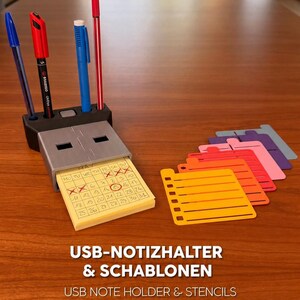 Op de afbeelding: Een grijze USB-vormige notitiehouder met een zwarte pennenstandaard voor vier pennen, met een geel mei-kalenderblokje. Daarnaast liggen vijf kleurrijke plastic sjablonen in geel, oranje, rood, roze en paars. De tekst luidt USB-NOTIZHALTER & SCHABLONEN.