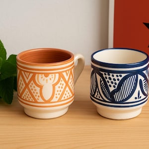 May include: Two ceramic mugs with detailed geometric designs. One is orange and cream, the other blue and white. The mugs sit on a wooden surface, with fresh mint and a red background with a black star.