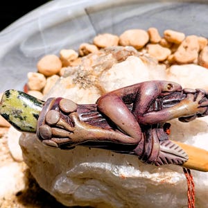 Peut inclure: Une sculpture détaillée et sculptée à la main d'une petite figurine nue allongée sur une base en bois. La figurine est de couleur brun violacé et la base est un mélange de brun et de beige. La sculpture repose sur un rocher blanc avec de petits galets.