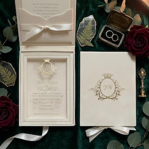 May include: A cream-colored wedding invitation set featuring a box with a white ribbon, a clear acrylic insert with gold text, and a matching cover with a gold crest. Includes two rings in a small box, dark red roses, and pressed fern leaves.
