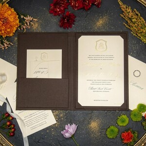 May include: A dark brown fabric folder containing a cream-colored wedding invitation with gold foil lettering and a crest. The invitation details the marriage of a daughter on February 24th, 2024, in New Orleans, Louisiana, with a matching RSVP card.