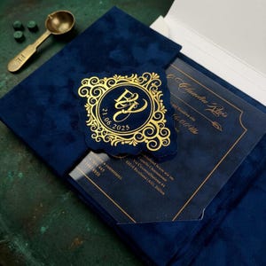 May include: A dark blue velvet invitation folder containing a clear acrylic card with gold text. A square navy blue wax seal with gold filigree and the initials E and J with the date 21.06.2025 sits on top. A brass wax melting spoon is nearby.