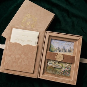 May include: A tan suede-textured presentation box containing wedding stationery. The left side features a pocket holding cards with the text Wedding Attire. The right side displays a card with a fountain illustration and a wax seal on a band.