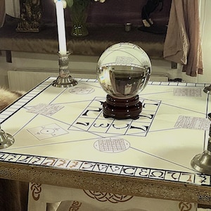 Pode incluir: Uma bola de cristal em um suporte de madeira repousa sobre uma mesa creme, adornada com padrões geométricos e números. Velas acesas em castiçais prateados adornam a mesa, criando um ambiente místico. A borda da mesa apresenta acabamentos decorativos.
