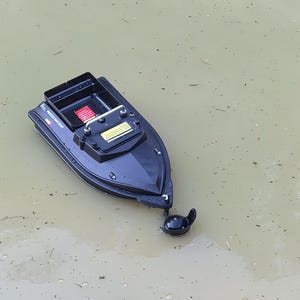 Könnte beinhalten: Ein schwarzes ferngesteuertes Futterboot schwimmt auf dem Wasser. Das Boot hat ein rechteckiges Fach, ein Bedienfeld und einen kleinen Propeller. Der Text "PROCESS BAIT BOAT" ist sichtbar.