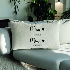 May include: A rectangular, light-coloured cushion with the words "Mom" and "Mimi" in black script, each with a heart symbol. The cushion also has the dates "-EST.1993-" and "-EST.2025-" printed below the names.