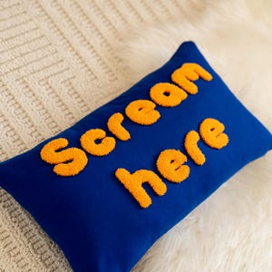 Könnte beinhalten: Ein rechteckiges blaues Kissen mit den Worten "Scream here" in orangefarbenen, strukturierten Buchstaben. Das Kissen liegt auf einer weißen, strukturierten Oberfläche.