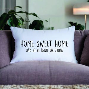 May include: White rectangular cushion with the text "HOME SWEET HOME" in black. Below the text is "OAK ST EL RENO, OK 73036". The cushion is on a purple sofa with a plant in the background.