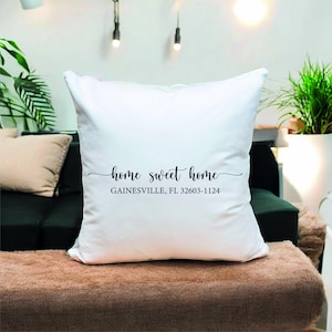 Può includere: Un cuscino quadrato bianco con la frase "home sweet home" in corsivo nero. Sotto la frase c'è il testo "GAINESVILLE, FL 32603-1124". Il cuscino è su una superficie marrone.