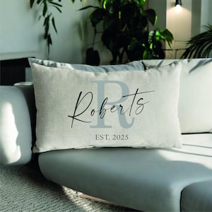 May include: A rectangular beige cushion featuring the name "Roberts" in black script and a large grey "R". Below the name is "EST. 2025". The cushion is on a grey sofa with other cushions.