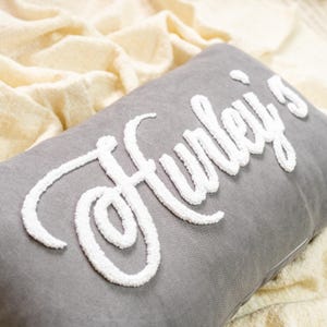 Peut inclure: Un coussin rectangulaire gris avec le nom "Hurley's" brodé en blanc, en écriture texturée. Le coussin repose sur un tissu crème texturé, créant une esthétique douce et accueillante. Article de décoration intérieure.