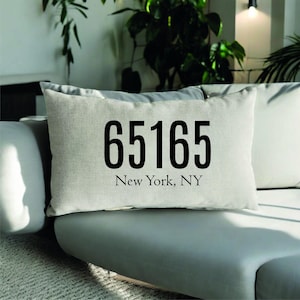 Może przedstawiać: Prostokątna, jasna poduszka z czarnymi cyframi "65165" i napisem "New York, NY". Poduszka leży na szarej kanapie, z innymi poduszkami i zielenią w tle.