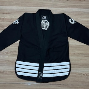 May include: Black martial arts uniform with white accents. The uniform features a collar, long sleeves, and a series of horizontal white stripes along the bottom. A circular logo is visible on the sleeves. The uniform is laid out on a wooden surface.