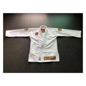 Puede incluir: Uniforme blanco de artes marciales con cuello y mangas largas, extendido sobre una superficie oscura. El uniforme tiene parches en las mangas y el pecho, y el texto "SHOYOROLL" es visible en la parte inferior.