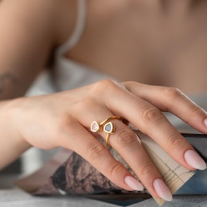 May include: A gold-colored ring with two gemstone accents, one pink and one dark blue, is displayed. The ring is worn on a hand with manicured nails, holding a magazine. The ring's design is open, with the gemstones set on either side of the finger.