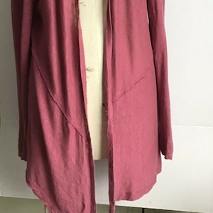 Pode incluir: Um cardigan de manga comprida, aberto na frente, em uma cor rosa empoeirado. O cardigan tem um caimento relaxado e uma bainha ligeiramente assimétrica.