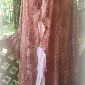May include: A long, dusty rose crochet cardigan with a delicate floral pattern. The cardigan has a shawl collar and long sleeves with bell cuffs. It is open at the front and has a flowing silhouette.