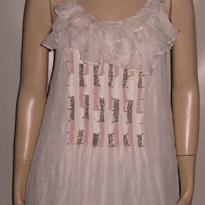 Puede incluir: Un vestido blanco sin mangas con un escote de volantes y un diseño de cuentas. El vestido tiene un dobladillo de volantes en capas.