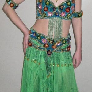 May include: A teal belly dance costume with colourful floral accents and beaded fringe. The outfit includes a halter top, armbands, a hip belt, and a flowing green skirt. The costume is designed for dance performances.