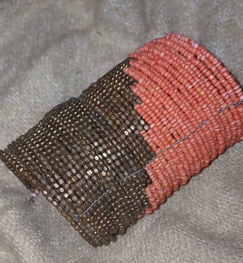 May include: A two-tone beaded bracelet. The bracelet is made up of bronze-coloured beads and coral-coloured beads. The beads are strung together with a thin, silver-coloured wire.