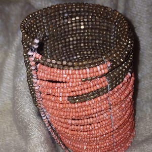 May include: A beaded cuff bracelet with a spiral design. The bracelet features rows of small, round beads in coral and bronze colours. The beads are strung together with thin wire, creating a flexible and textured accessory.
