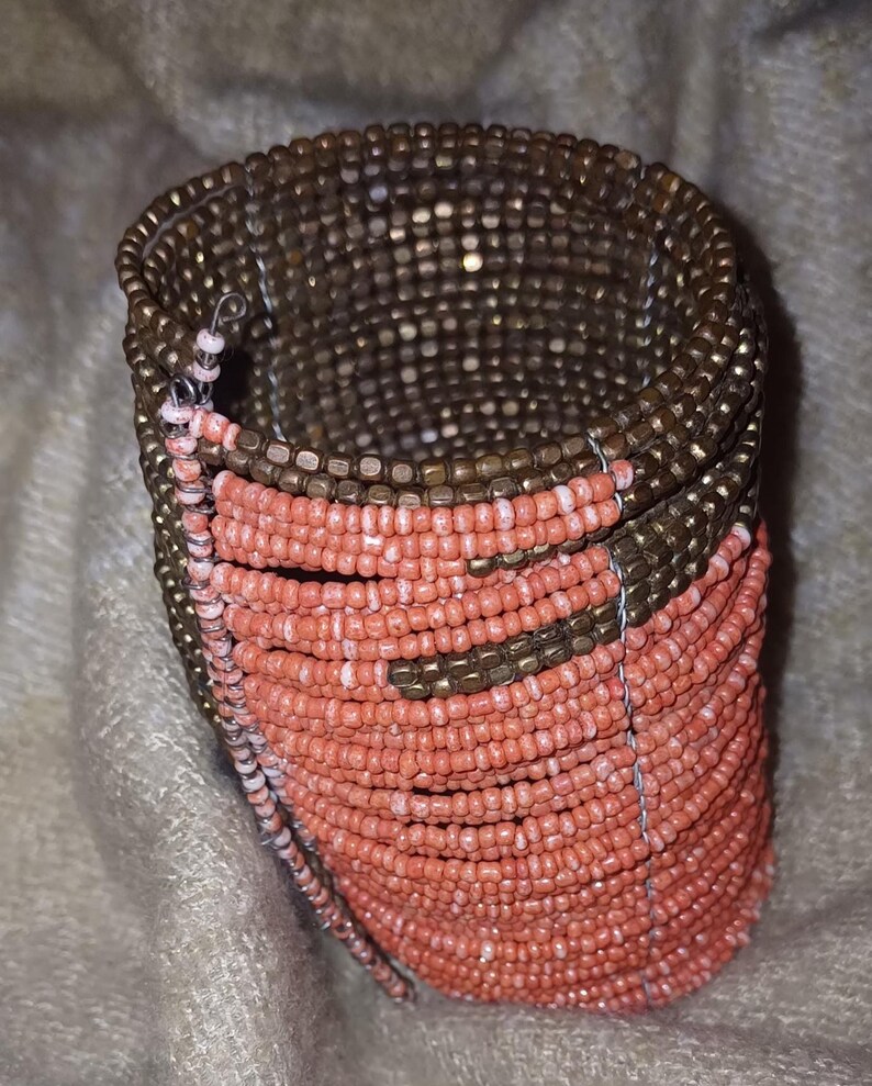 May include: A beaded cuff bracelet with a spiral design. The bracelet features rows of small, round beads in coral and bronze colours. The beads are strung together with thin wire, creating a flexible and textured accessory.