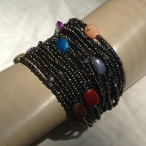 Può includere: Un braccialetto di perline nere con pietre colorate. Il braccialetto è fatto di molti fili di piccole perline nere e presenta diverse pietre rotonde di vari colori, tra cui blu, rosso, rosa e viola.