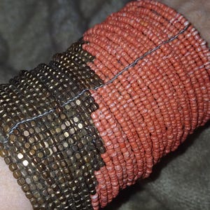 May include: A two-tone beaded cuff bracelet. The bracelet is made of small, round beads in bronze and coral hues. The beads are strung together with a thin wire, creating a textured surface.