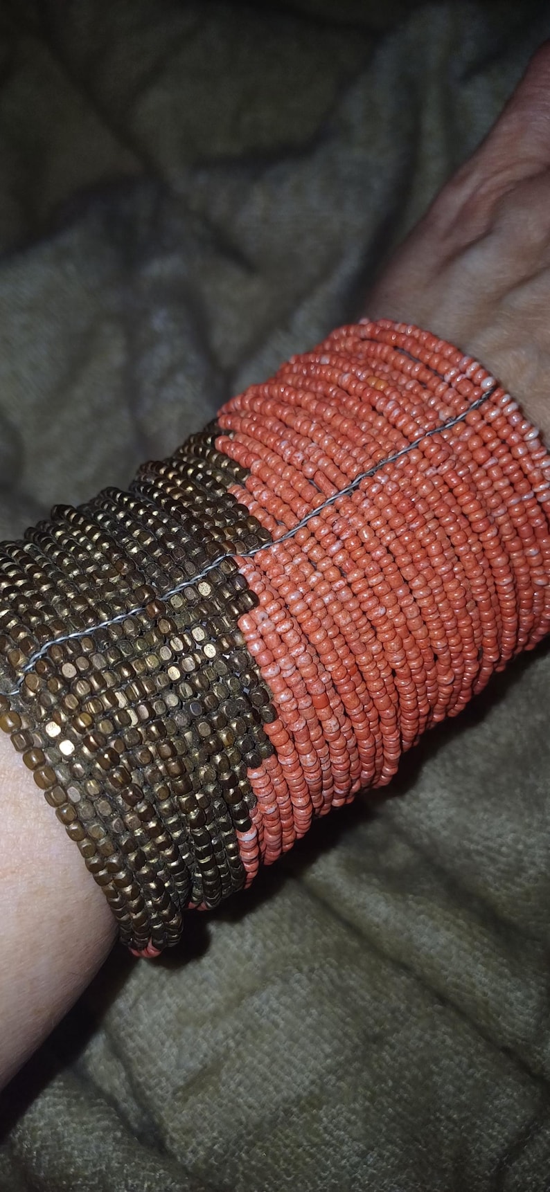 May include: A two-tone beaded cuff bracelet. The bracelet is made of small, round beads in bronze and coral hues. The beads are strung together with a thin wire, creating a textured surface.