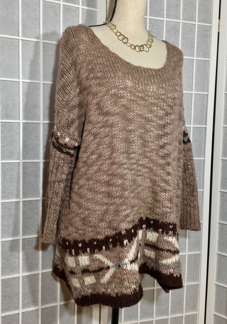 May include: A brown knit sweater with a scoop neck and long sleeves. The sweater features a patterned design in shades of brown, white, and teal at the hem and cuffs. A gold-colored necklace is visible around the mannequin's neck.