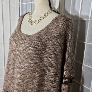 May include: A brown, open-weave sweater with a scoop neck and long sleeves. The sweater is displayed on a mannequin and paired with a silver chain necklace. The sleeves have a patterned design near the cuffs.